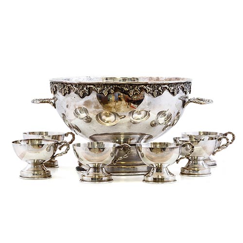 Sub.:22 - Lote: 1270 -  Ponchera y seis tacitas en metal plateado, con asas cincelada y la ponchera con racimos de vid en el borde.